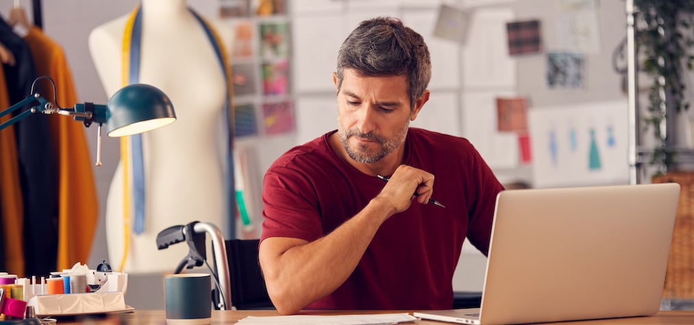 Mature Male Fashion Designer In Wheelchair Sitting At Desk In Studio Working On Laptop